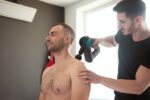 A physiotherapist provides shoulder massage therapy with a massage gun in a clinic.
