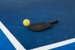 A pickleball paddle and yellow ball on a blue court corner, symbolizing sport and activity.