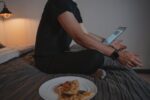 Person using laptop on bed with pizza, depicting a cozy home office setup.