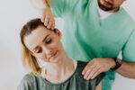 Serious doctor in uniform massaging smiling female patients neck and stretching stiff neck muscles against gray background